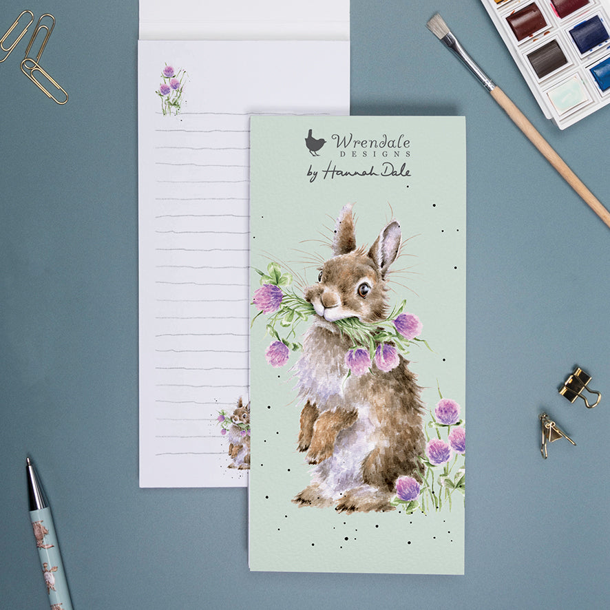 shopping list notepad with pale green front cover and illustration of a rabbit holding a bunch of flowers. there is a wrendale designs logo at the top.  The open notebook shows mini illustrations at the bottom of each lined page.