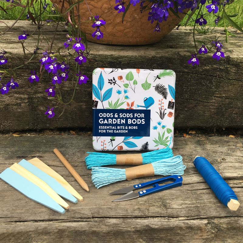 tin with essential bits and bobs for the garden printed on a floral design and contents of tin displayed in front.  the tin is sitting in a garden scene. The contents are mini snips, pencil, wooden labels and gardening rafia and twine