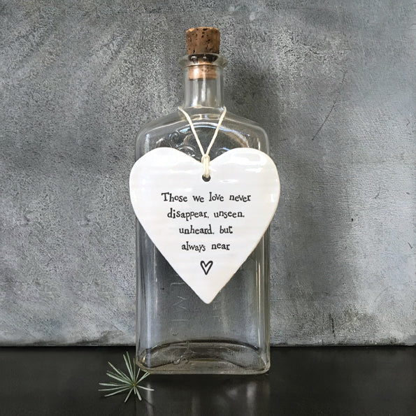 white porcelain heart with twisted paper cord hanging loop.  The words Those we love never disappear, unseen, unheard, but always near are on the front with a little heart underneath displayed hanging on a glass bottle