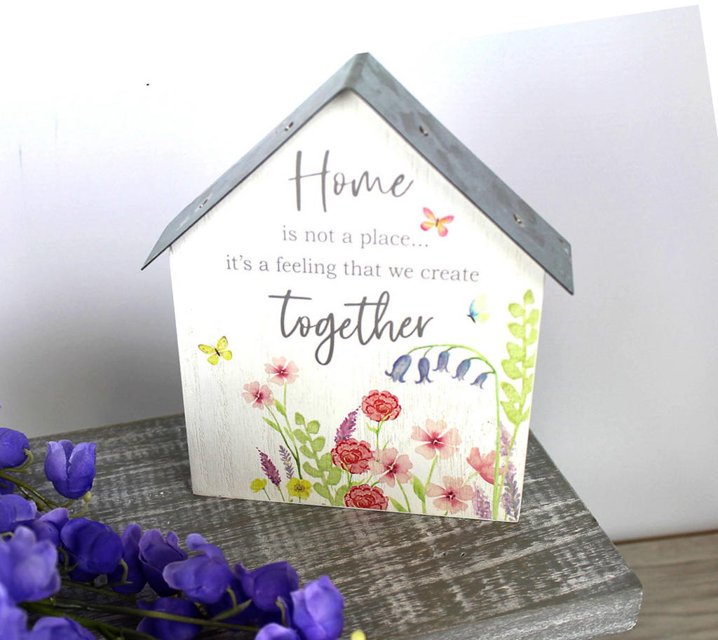 wooden house shaped free-standing shelf sitter with words home is not a place ...it's a feeling that we create together.  The sign is white painted wood and  has a floral colourful deisgn